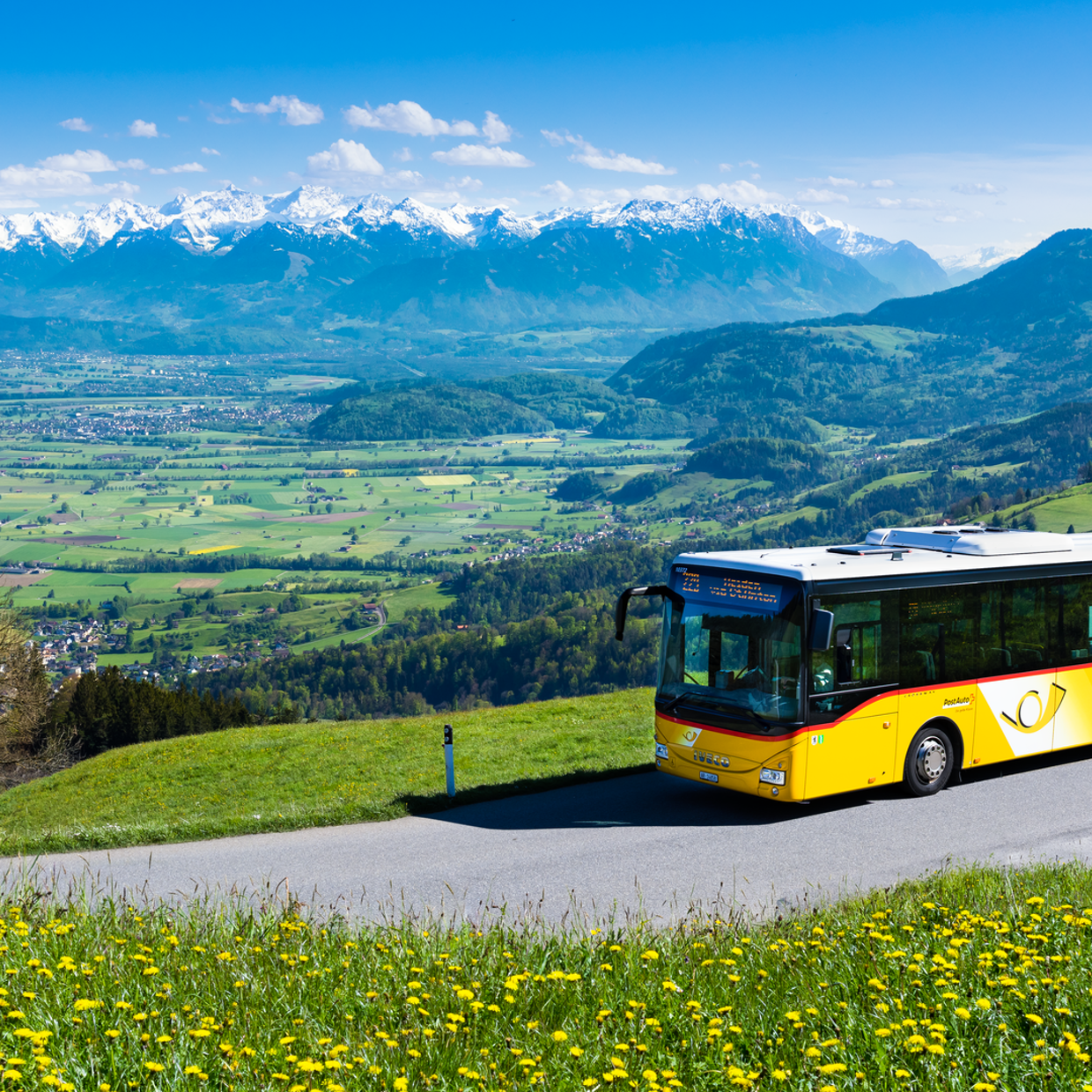 Ein Postauto auf einer Landstrasse mit grossen Wiesen und Bergen im Hintergrund